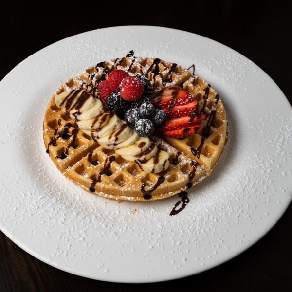 Waffle topped with sliced bananas, mixed berries, chocolate drizzle, and powdered sugar on a white plate.